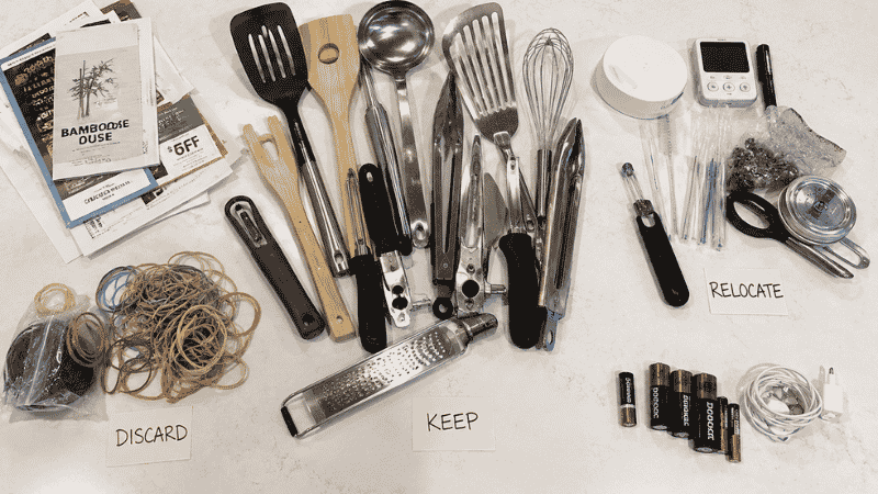 Kitchen drawer contents spread on counter sorted into keep, relocate and discard piles