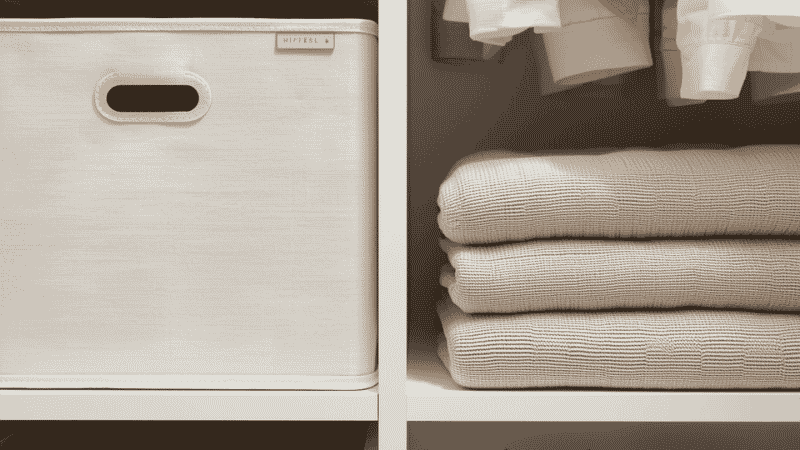 A close-up of organized closet shelving featuring high-end linen-textured storage bins and neatly stacked neutral cashmere sweaters.