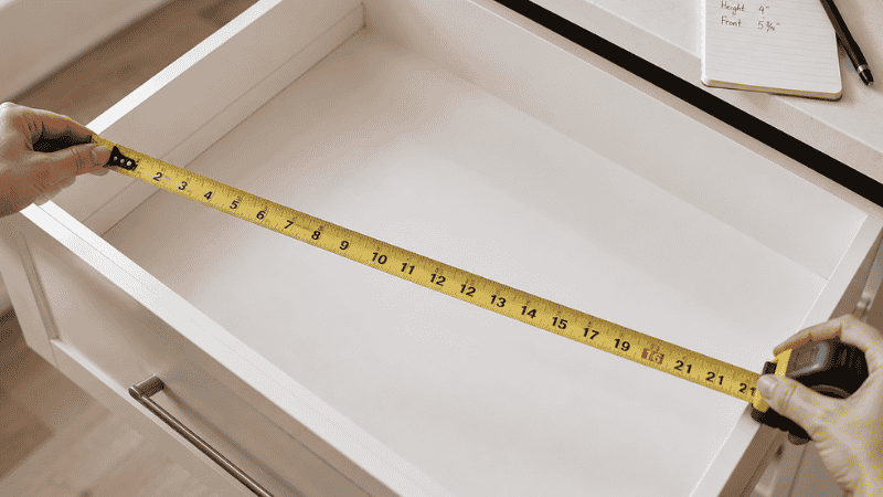 Hands measuring the interior width of an empty kitchen drawer with a tape measure