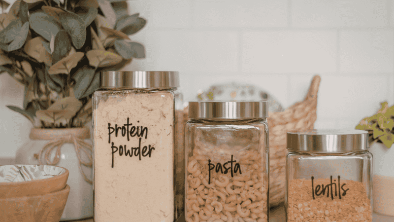 A curated pantry interior with uniform glass jars, minimalist labels, and acacia wood lids, complemented by woven seagrass baskets for a warm, Organic Modern storage aesthetic.