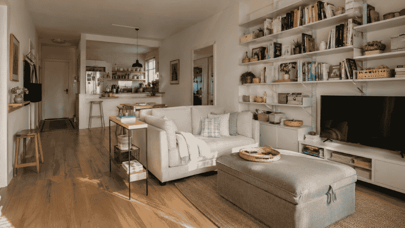 Small apartment living room with storage ottoman tall shelving and organized open kitchen in background