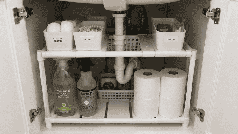 Under bathroom sink cabinet organized with expandable two-tier shelf unit around plumbing pipes