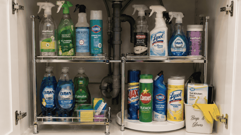 Under kitchen sink cabinet organized with two-tier shelf unit around plumbing turntable for bottles and labeled bin