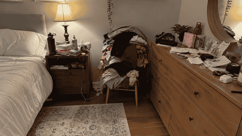 Cluttered bedroom showing chair covered in clothes pile and overcrowded dresser surface before organizing
