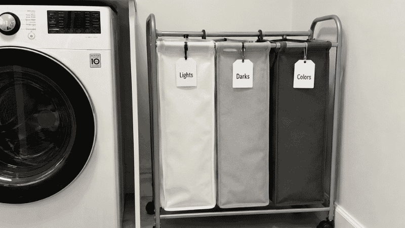 Three compartment rolling laundry sorter with lights darks and colors labels beside washing machine