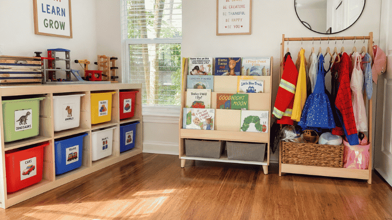 Organized playroom showing labeled toy bins at child height forward facing bookshelf and dress up rack with clear floor