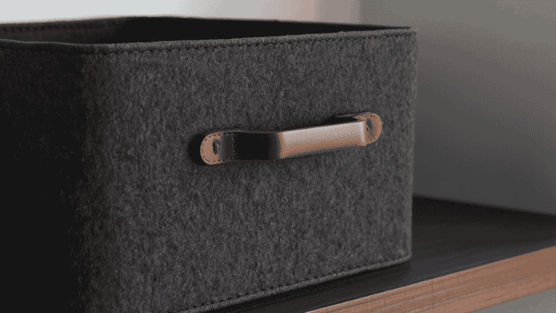 A close-up of the charcoal grey felt storage box, featuring a minimalist brown leather handle, is placed gracefully on a dark walnut shelf, showcasing the high-end texture and craftsmanship details created by the combination of different materials.