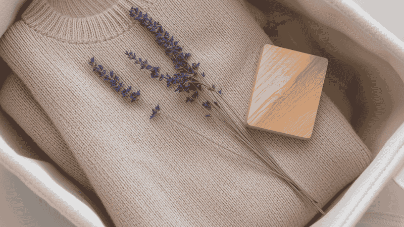 Close-up shots of the daily life details inside the canvas storage box: A neatly folded cashmere sweater, next to it is a small bunch of dried lavender and a piece of cedar wood, conveying a refined lifestyle that is natural and cares for the fabric.